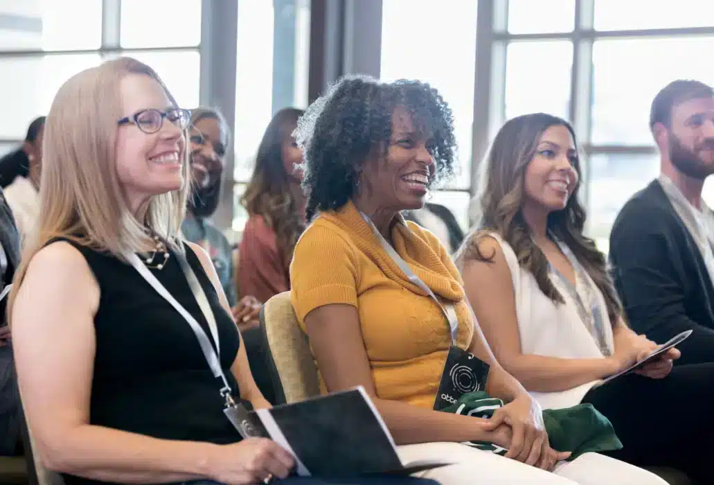 Smiling group of diverse adults seated in a bright conference room with lanyards, listening to a presentation