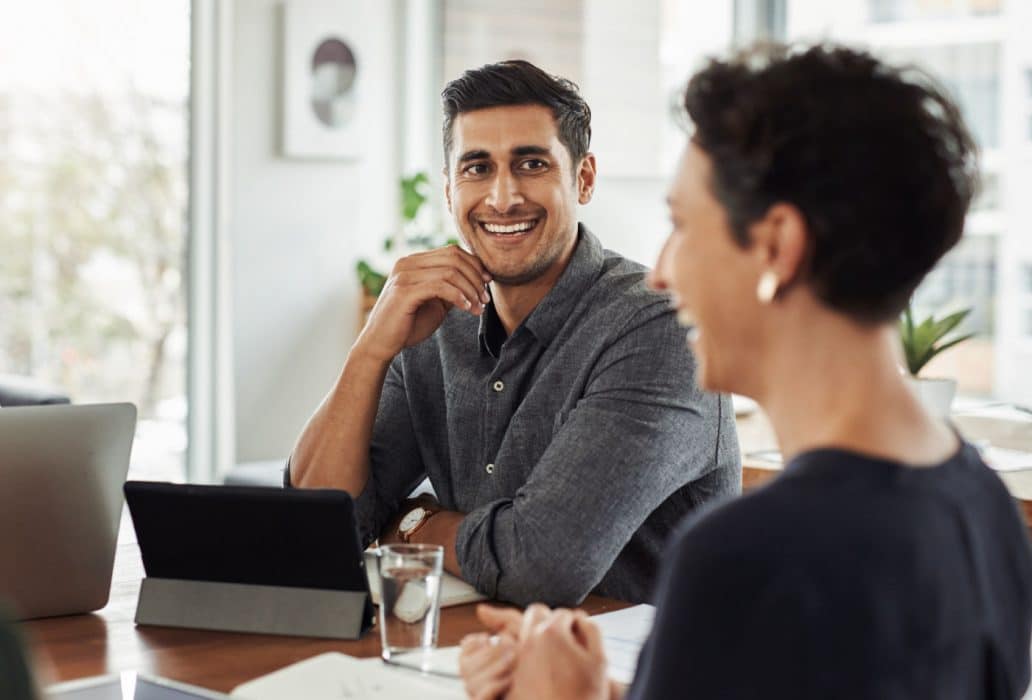 A man and a woman sitting at a table in an office setting and laughing while working together.