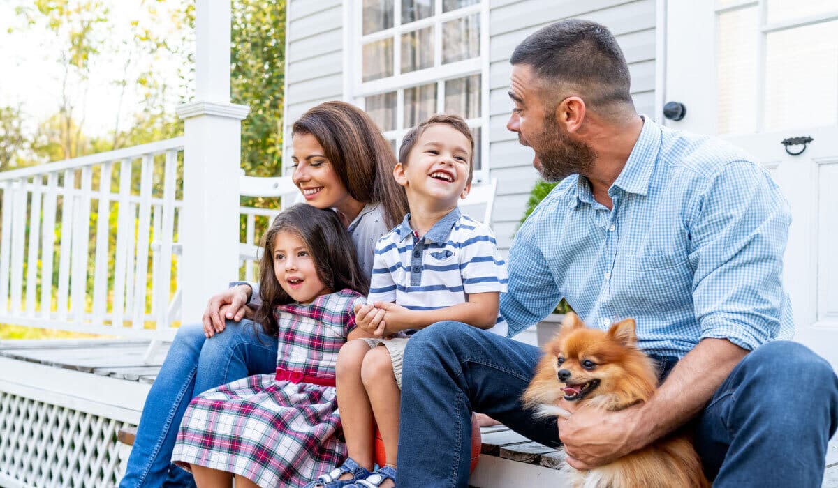 A family sitting on the front porch of a house, there's a dog sitting with them and they are laughing.