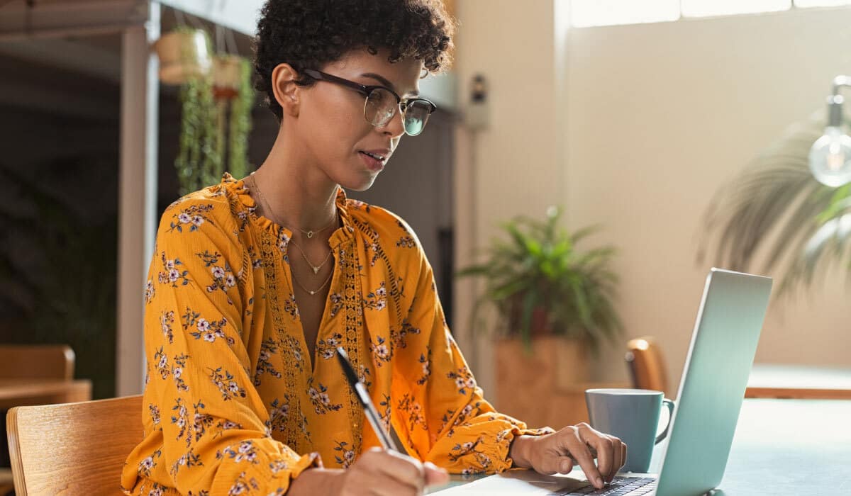 A woman sitting at a table and using a laptop and writing something down.