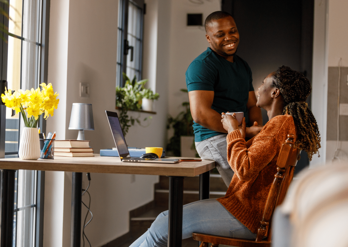 Couple talks at a kitchen table while one drinks coffee and has the laptop open