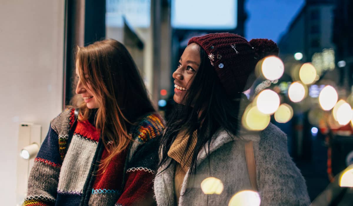 Two women looking into a store window and laughing, there are holiday lights in the background.