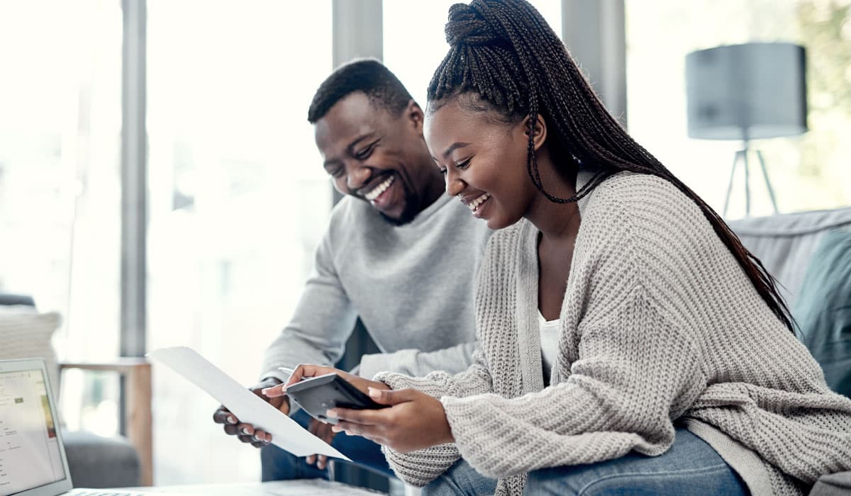 A couple sitting on a couch looking at a piece of paper and laughing.
