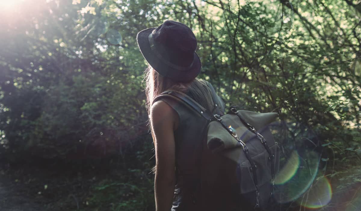 a woman walking through a forest. She is wearing a backpack and a hat.