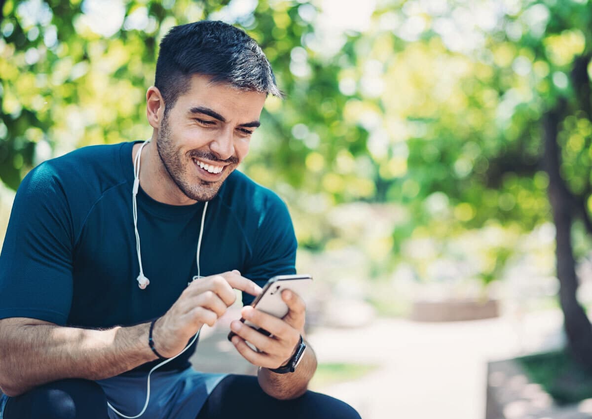 A man sitting outside and smiling at a cell phone, he has headphones around his neck.