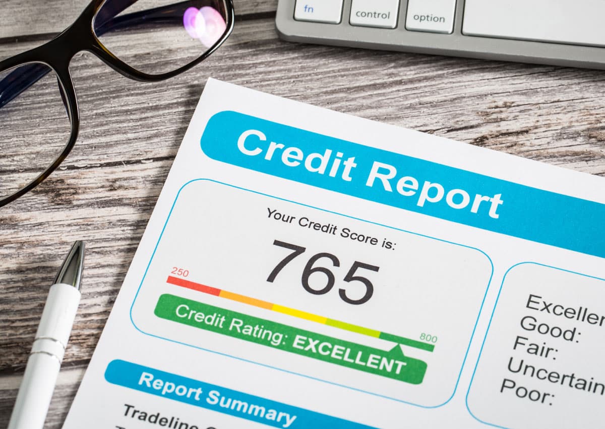 Paper titled Credit Report and displaying a 765 credit score lays on a desk with a pen, glasses, and the bottom left of a keyboard.