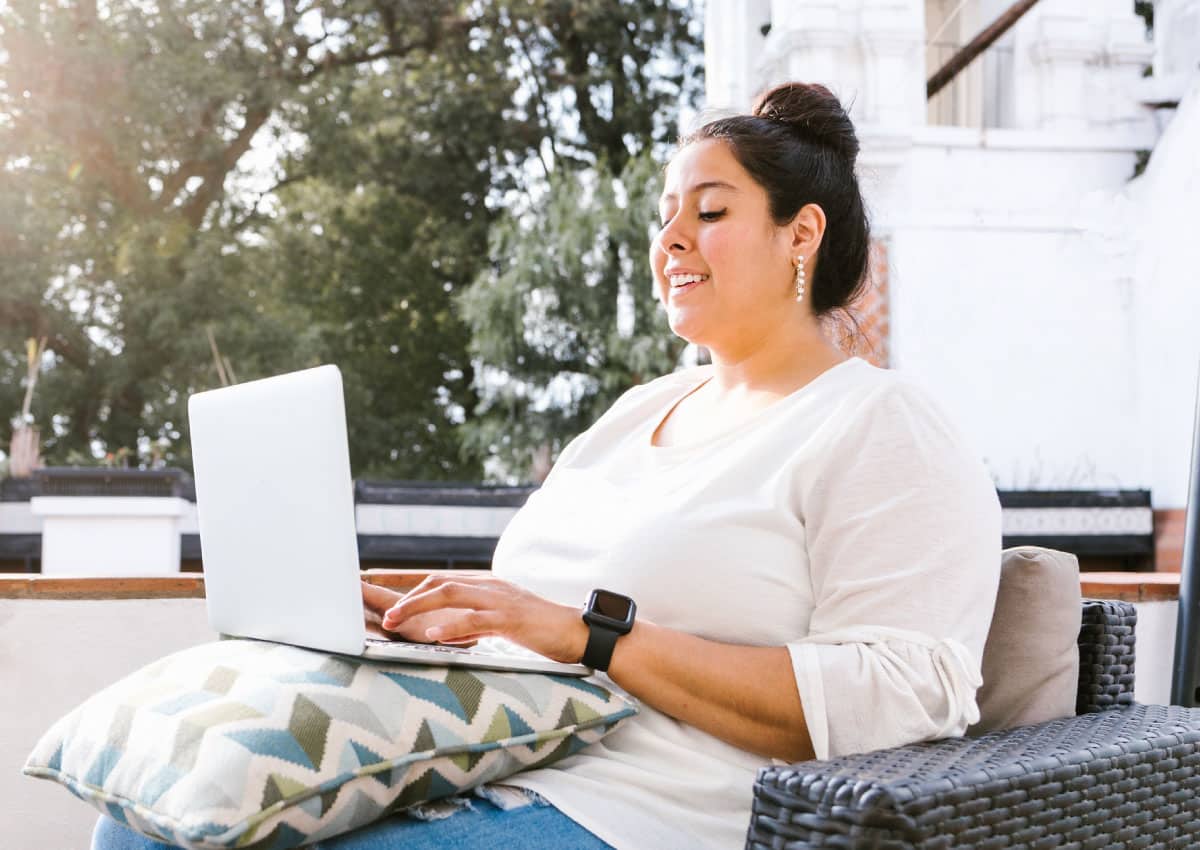 A woman sitting on outdoor furniture using an open laptop.