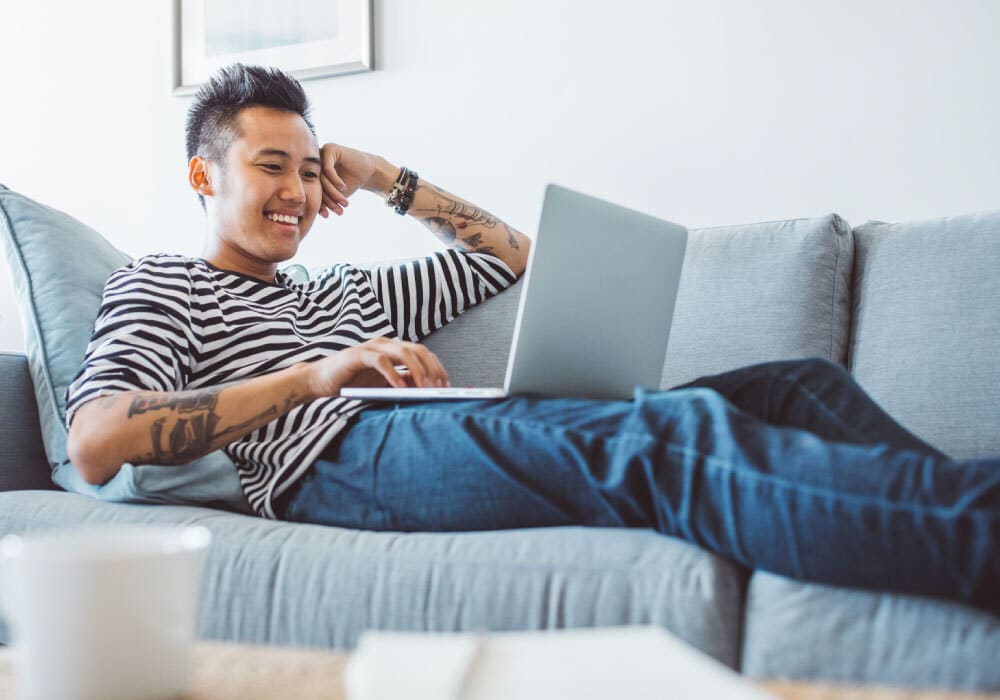 Man sitting on a couch with a laptop on his lap. He is smiling at the computer.