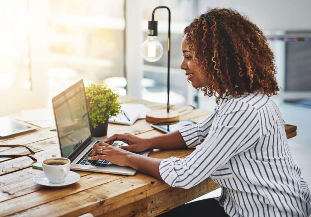 Woman sits at wood table typing on laptop.