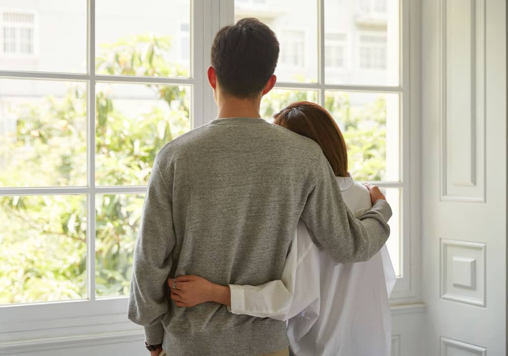 Two people stand looking out the window, one has their arm around the other.