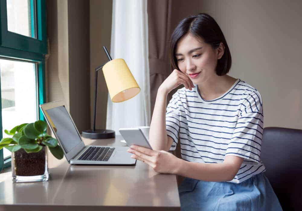 Woman sitting at a desk looking at her phone with an open laptop next to her