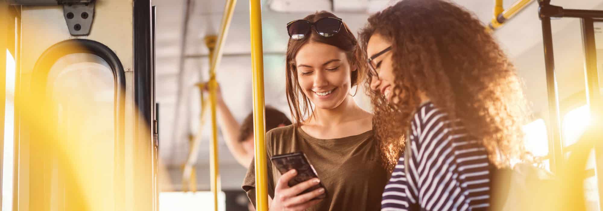 Two girls stand on public transportation and are smiling down at a phone.
