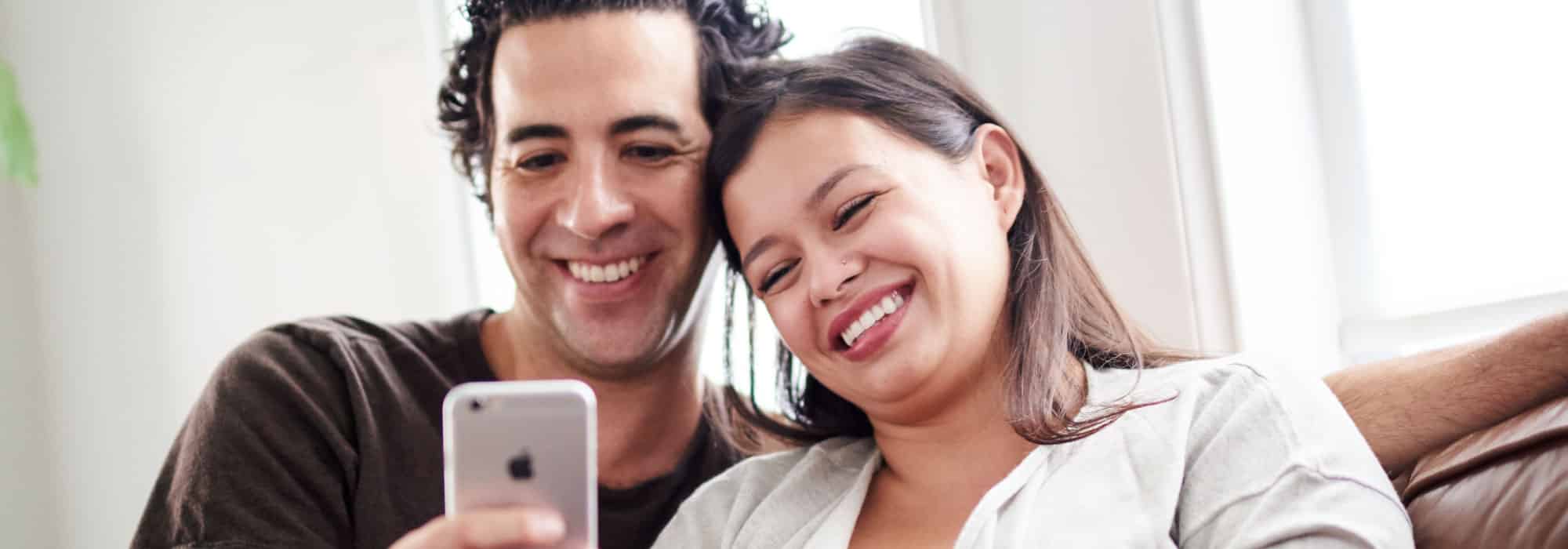 A couple sits on a couch and looks at the screen of a cell phone together. They are laughing.