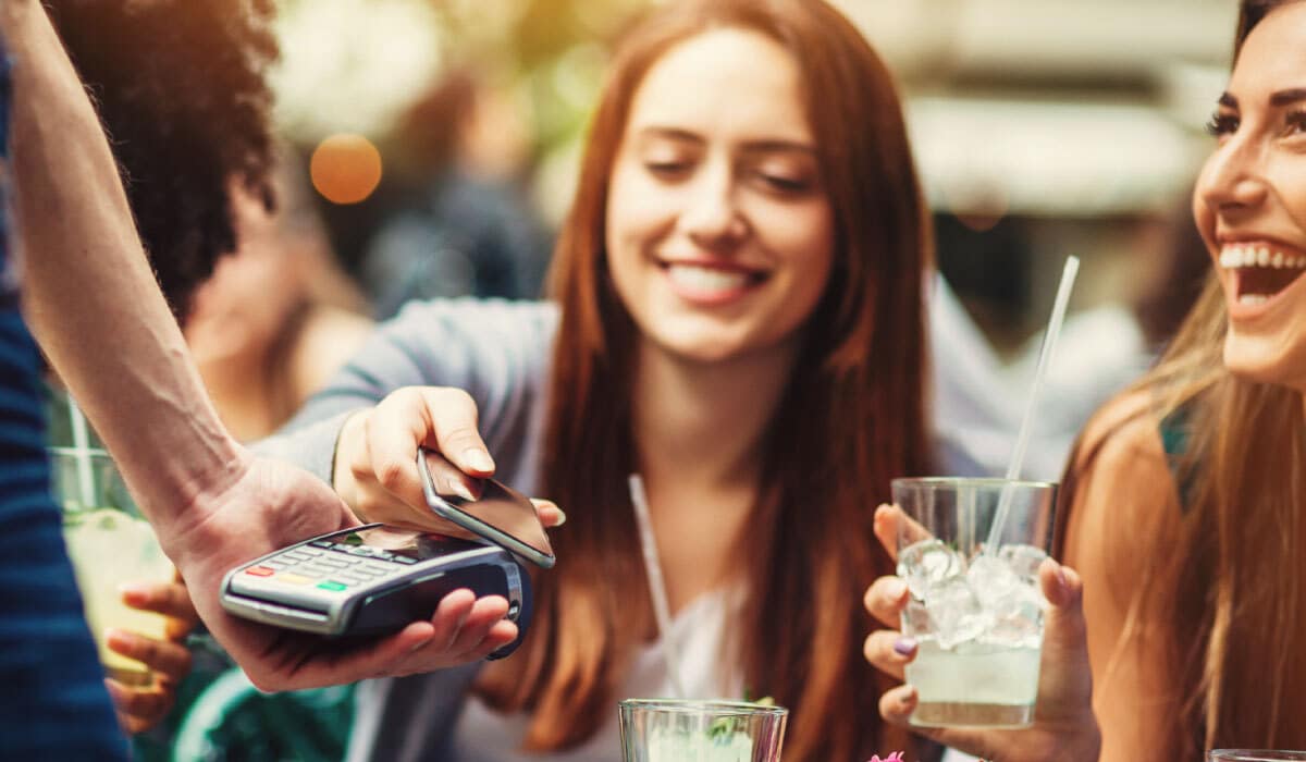 Two girls paying for drinks by using their phone and a credit card reader. They are smiling and laughing.