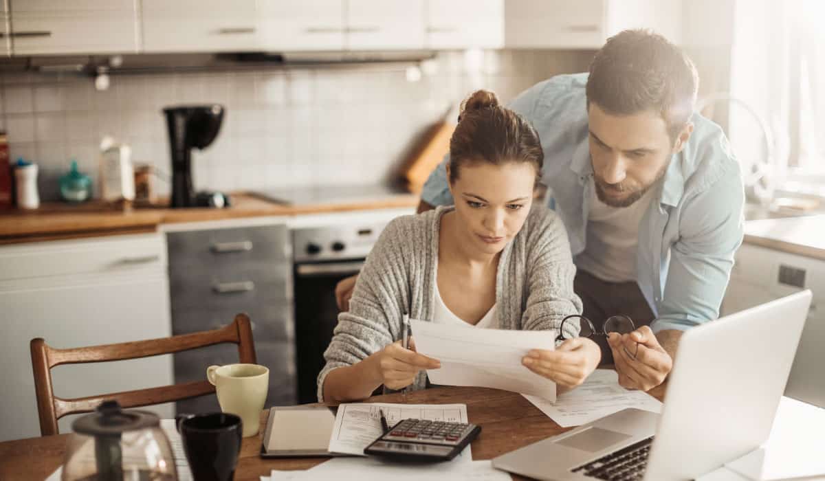 A couple sits at a kitchen table, they are looking at bank statements and there is a laptop open in front of them.