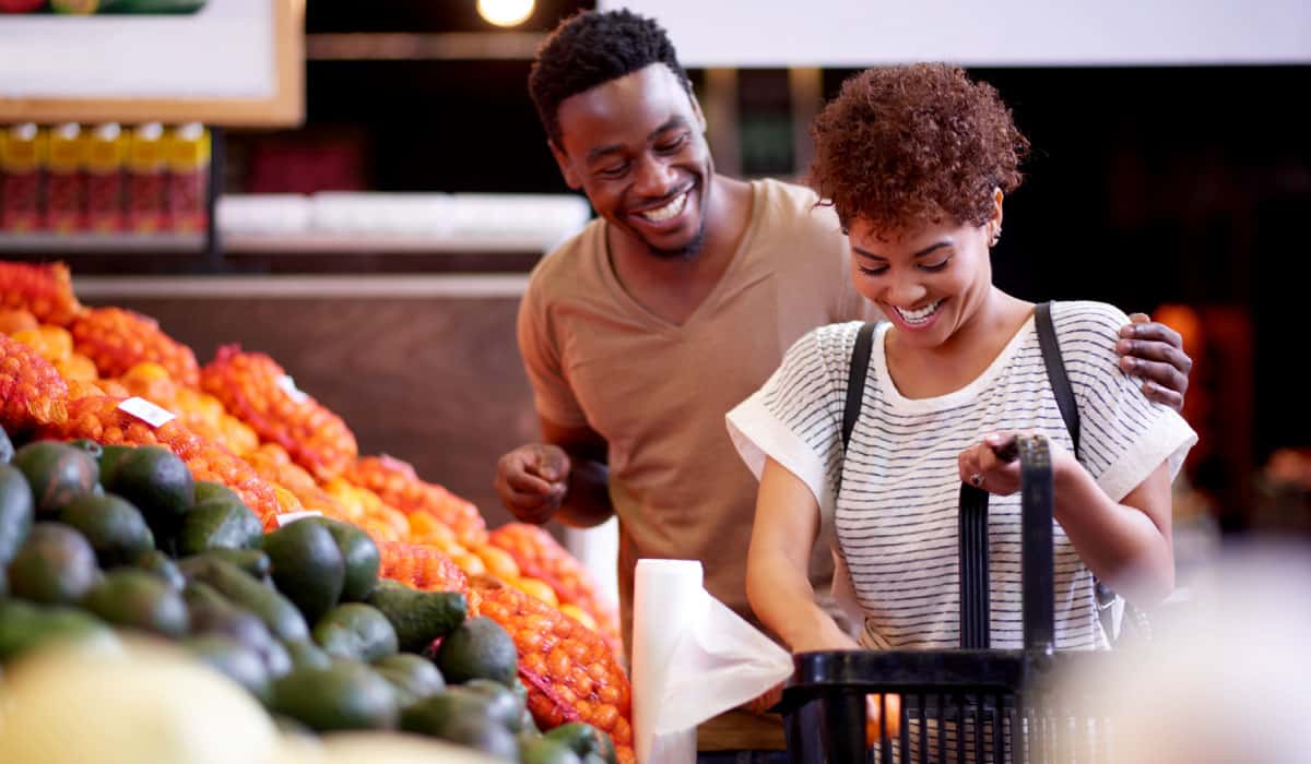 A couple shopping at the grocery store. They are standing in front of produce. The woman puts some produce into a basket and they are laughing.