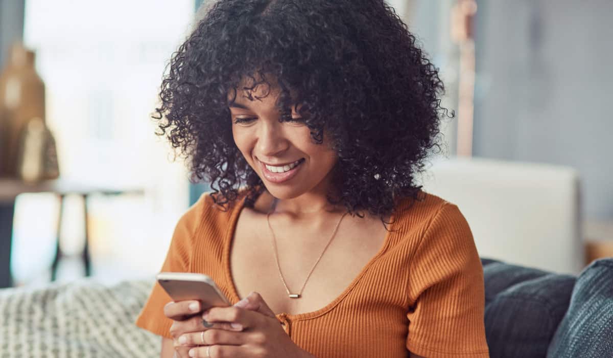 A woman sitting on a couch and looking down at a cell phone, she is smiling.