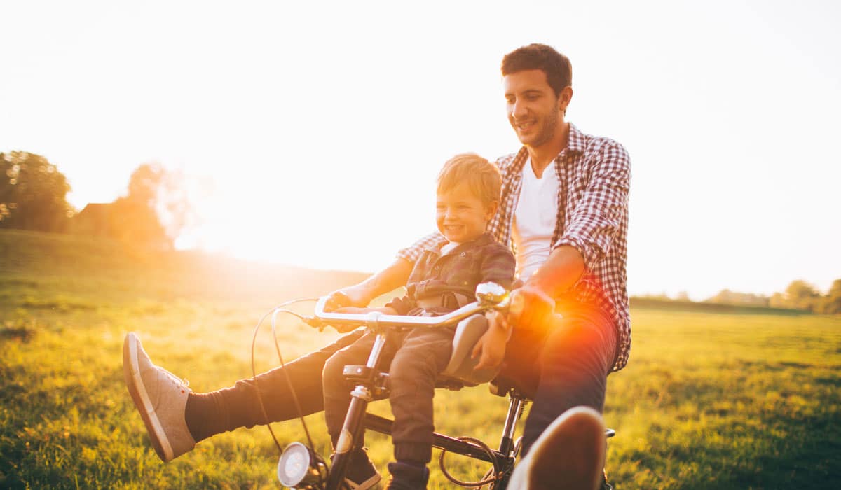 A man riding a bike in a field. There is a little boy sitting on the handlebars. They are smiling and laughing.