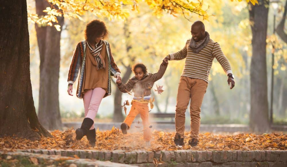 A family walking through a forest, it appears to be fall, there is a young child between them. Each one of them is holding one of the child's hands, and the child is between them.