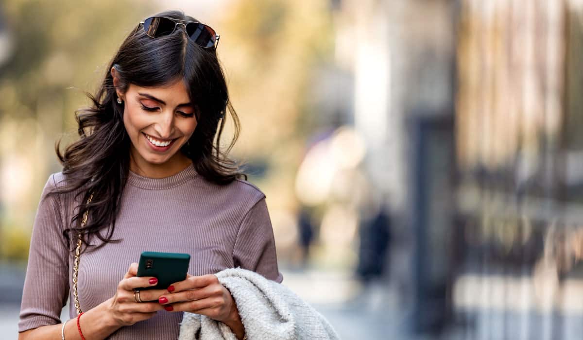 Woman walking and looking down at a cell phone, she is smiling.