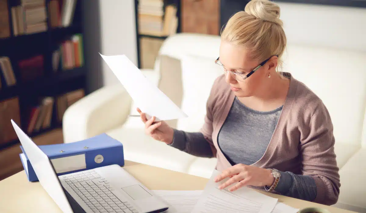 Woman sits at table with laptop open while looking at papers.