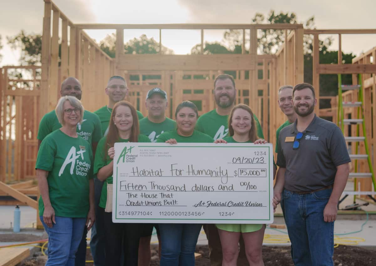 A+FCU volunteers present a check for $15,000 to Habitat for Humanity and pose in front of a framed out house.