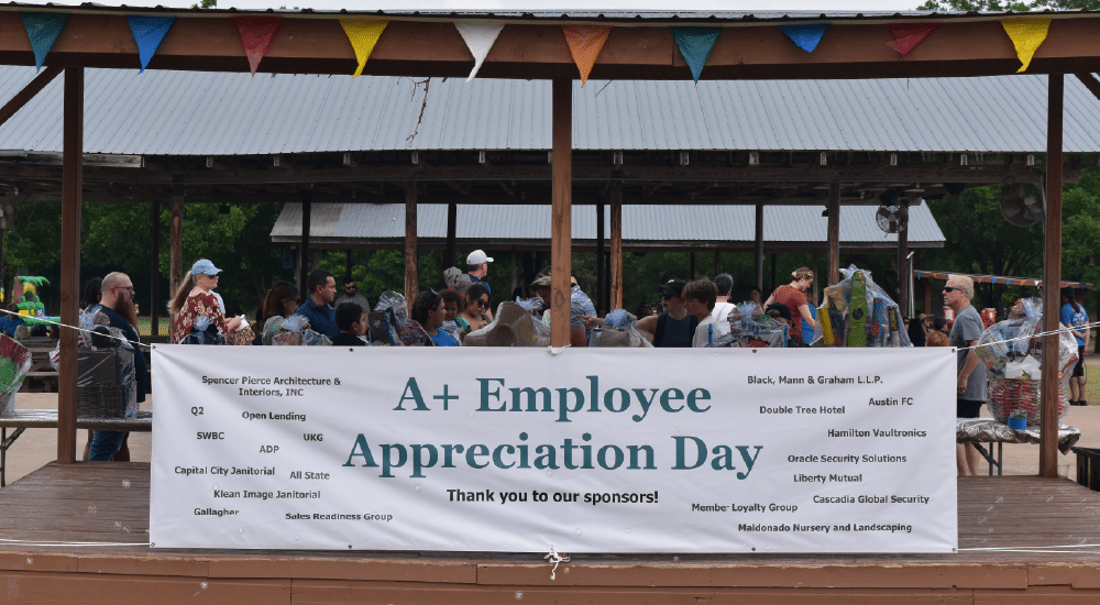 Giant white sign that says A+ Employee Appreciation Day with a list of sponsors hangs outside in front of group of employees