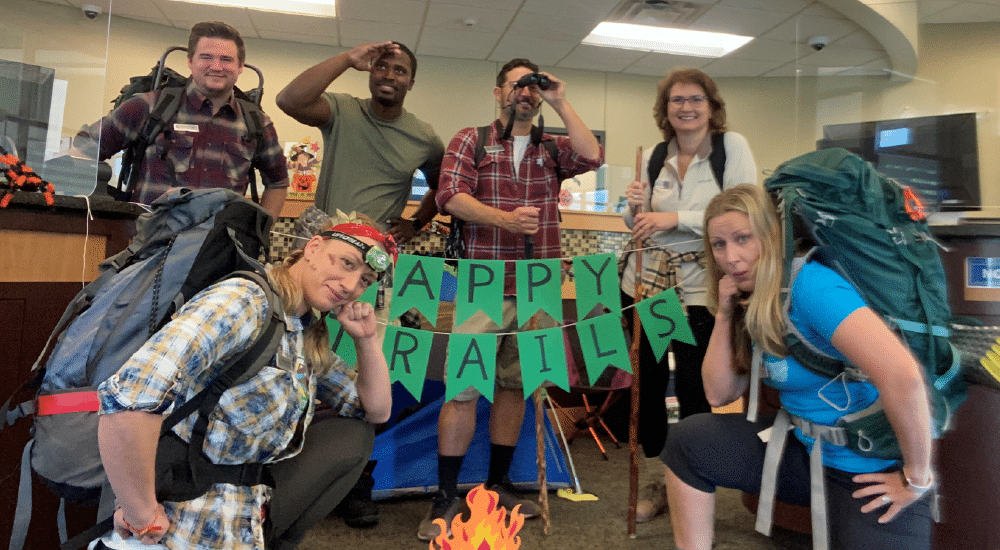 A+FCU team members in camping attire pose in front of a fake campfire and sign that says Happy Trails for Halloween.