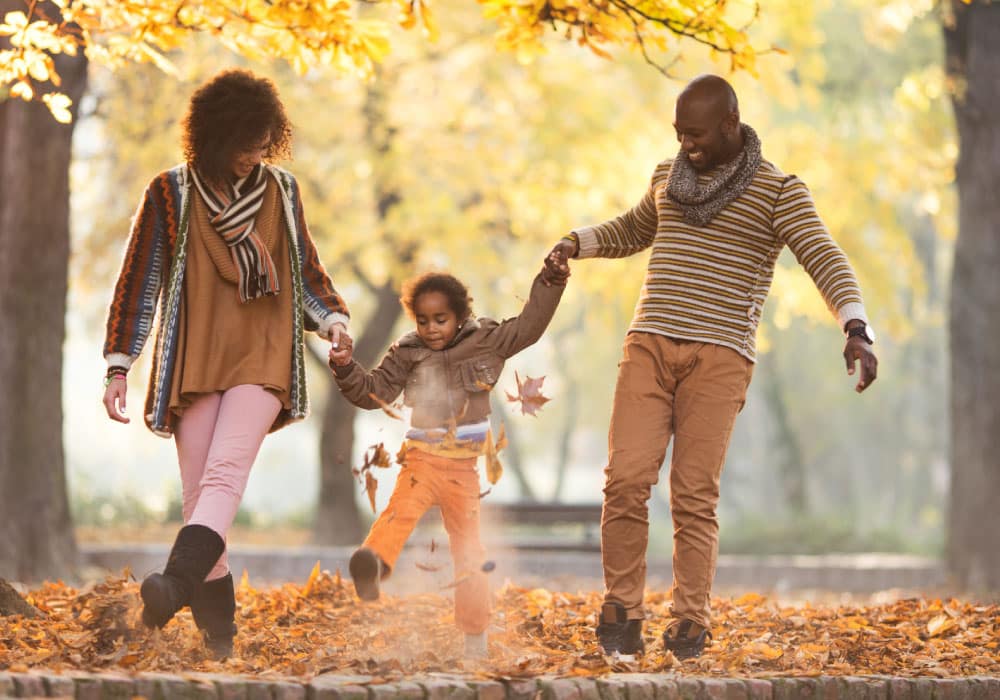 A family walking through a forest, it appears to be fall, there is a young child between them. Each one of them is holding one of the child's hands, and the child is between them.