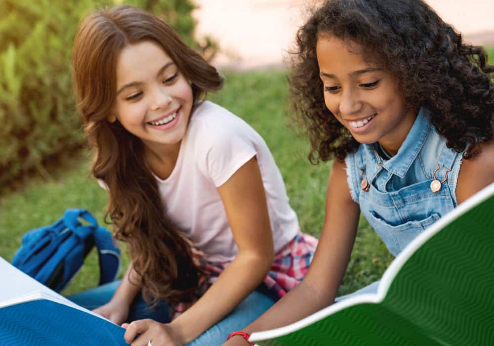 Two girls sitting on grass and sharing books, they are laughing and smiling.