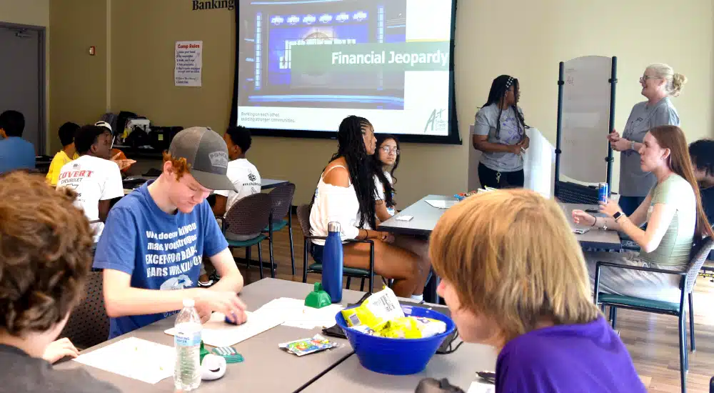 Students sit at tables in a classroom, watching a projector screen that reads Financial Jeopardy.