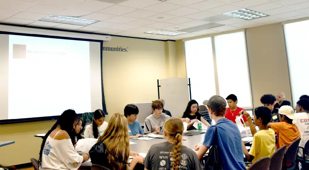 Group of students around a conference table in a classroom, actively discussing with a projector screen at the front.