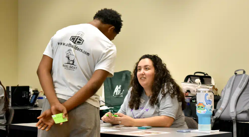 Man in a white T-shirt hands sticky notes to a seated woman at a table during a workshop session.