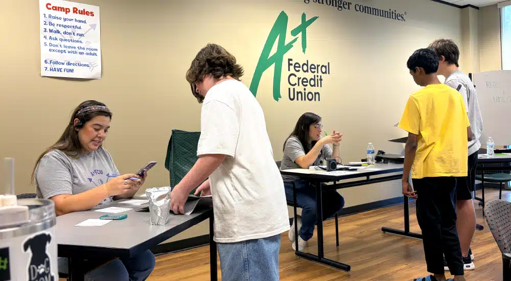 People receive help at a community service desk inside a credit union; staff assist customers under a large A+ Federal Credit Union sign on the wall.