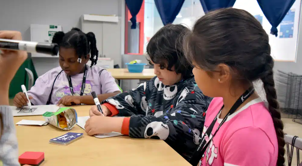 Three children sit at a classroom table drawing with markers; pink-shirt girl with a braid on the right.