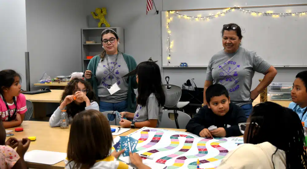 Group of children around a long table playing a colorful board game; two teachers stand nearby in a classroom with string lights.