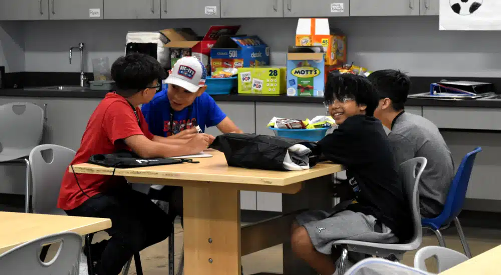 Four boys sit around a table in a classroom-like kitchen area, collaborating on a project with snacks on the counter behind them.