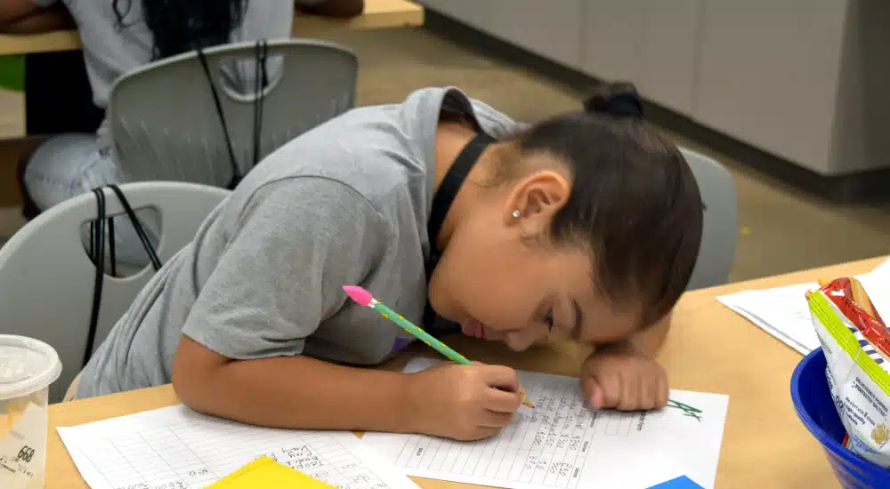 Girl leaning over a desk, writing with a pink pencil on a worksheet in a classroom setting.