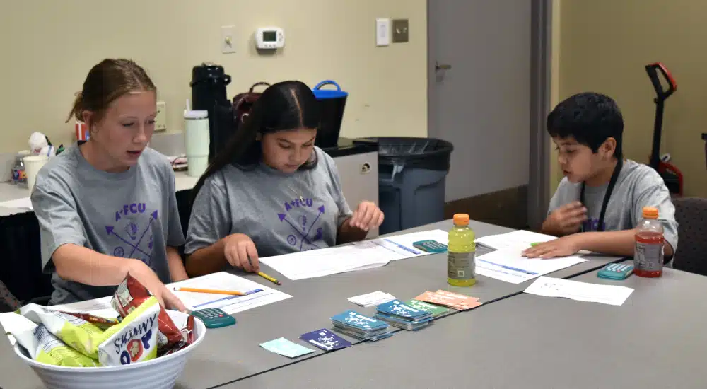 Three children sit at a classroom table, working on worksheets with pencils and cards laid out between them.