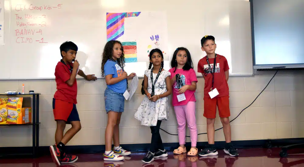Five children stand in a classroom in front of a whiteboard with a colorful poster.