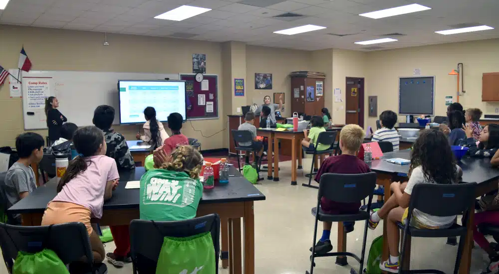 Teacher leads a lesson at the front of a classroom, presenting on a large screen while students sit at lab tables watching.