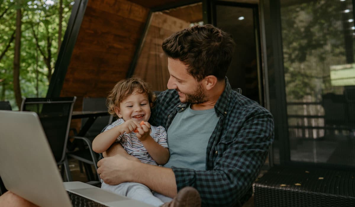 A man sitting on a porch with a child on his lap. There is a laptop in front of them.